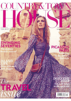 Woman in a flowing purple patterned dress with a red crossbody bag standing by a rustic stone archway