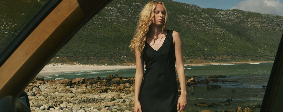 Sleeveless black dress in a relaxed silhouette worn by woman standing by rocky shoreline, framed through a car window