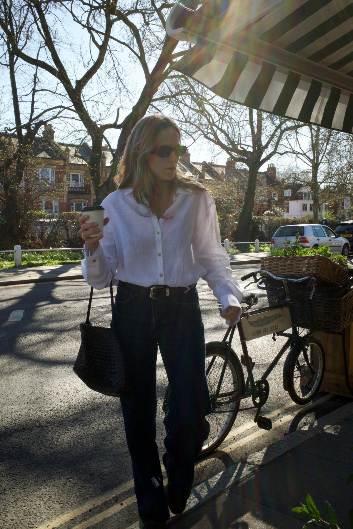 Woman in a loose white shirt and dark jeans walking on a sunlit street holding a coffee cup and black woven handbag