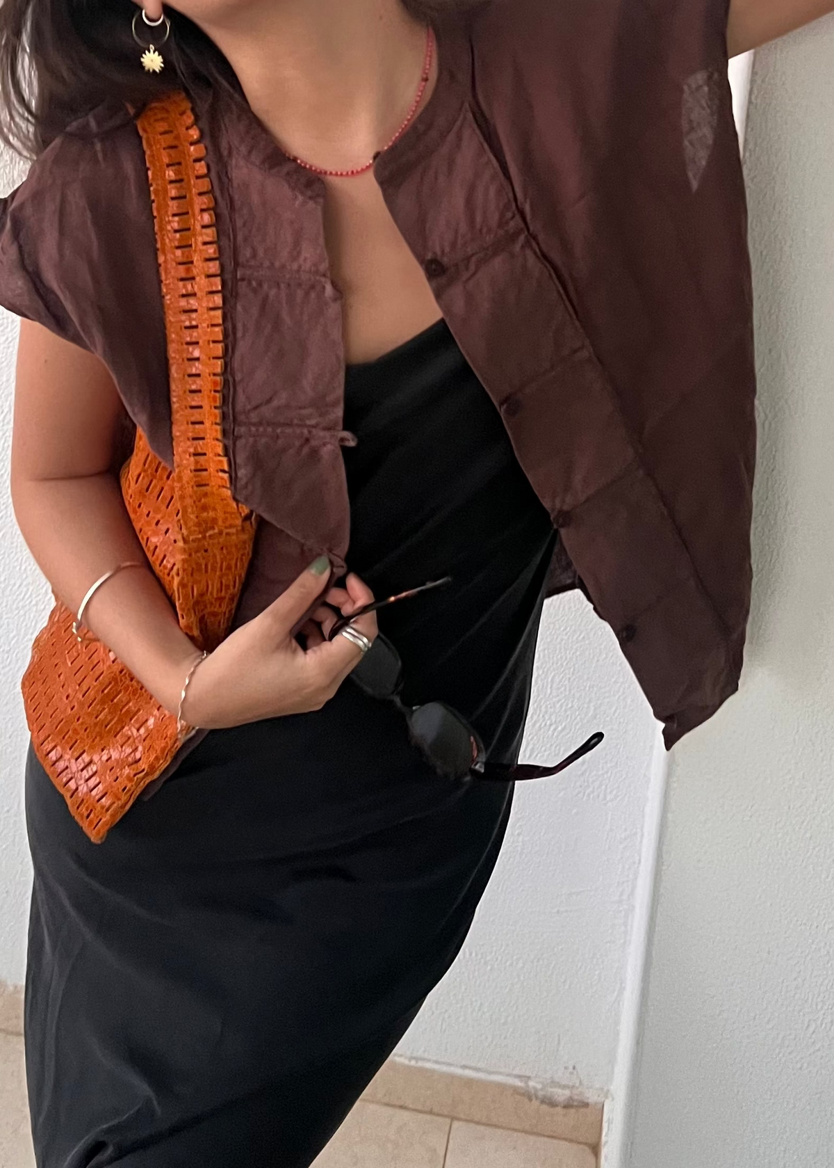 Close-up of a person in a brown shirt over a black dress, holding black sunglasses and an orange woven bag with a gold sun earring