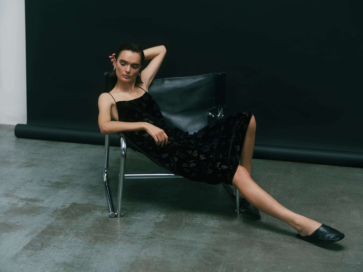 Woman reclining in a black leather and chrome chair wearing a sleeveless black dress with floral pattern and black slip-on shoes