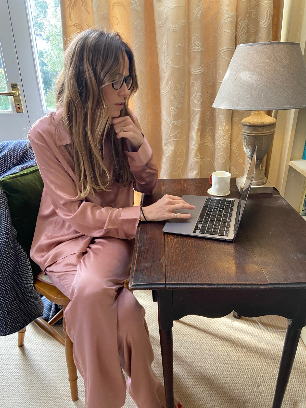 Woman sitting at a dark wooden table wearing a soft pink satin pajama set working on a laptop in a softly lit room