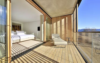 Balcony with wooden floor and slatted walls, a white cushioned lounge chair, side table, and mountain view under blue sky