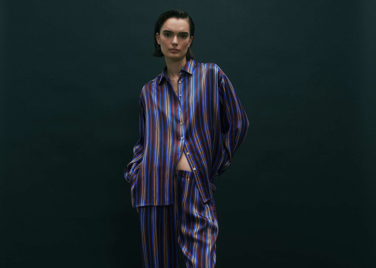 Model wearing loose-fitting silk pyjama set featuring vertical stripes in blue, purple, red and white on dark background