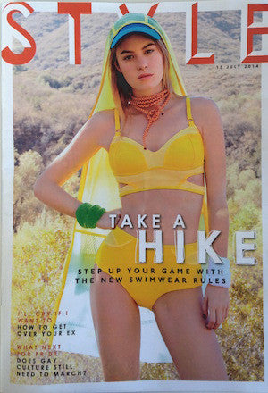 Model in a bright yellow structured bikini with belt detail, green cap with yellow hood, and orange beaded necklace outdoors