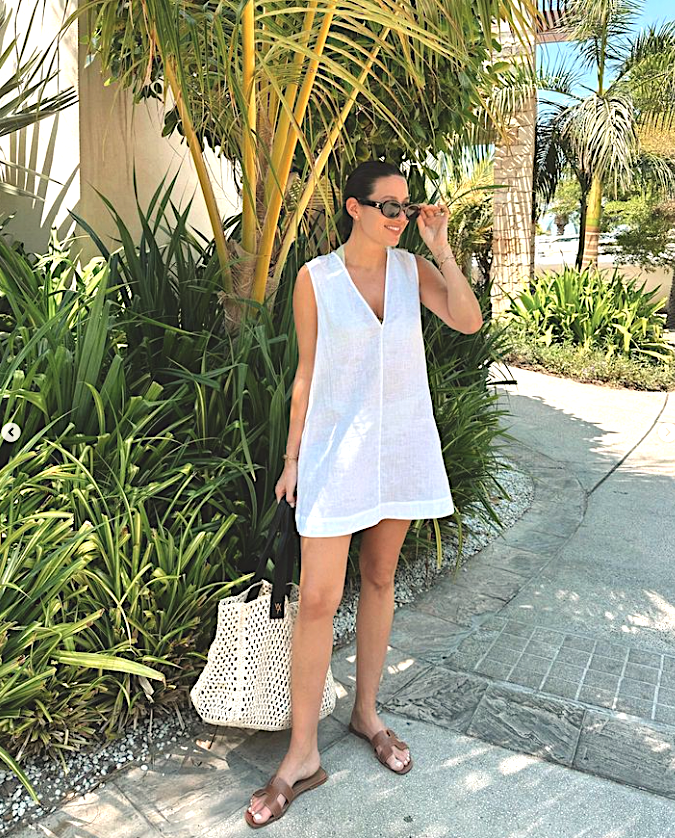 Woman outdoors in a sleeveless white linen dress with brown sandals, holding a large white woven tote with black handles