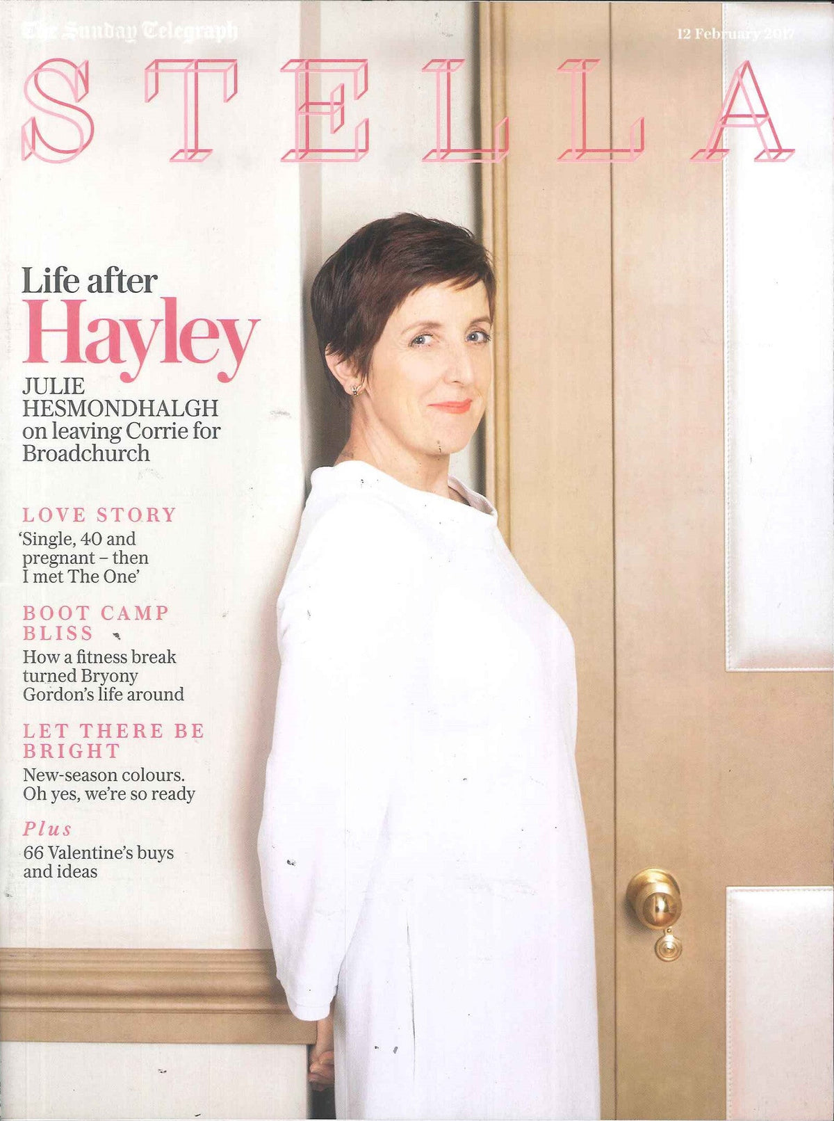 Woman with short dark hair in a loose white top standing sideways against a cream wall and wooden door with brass handle