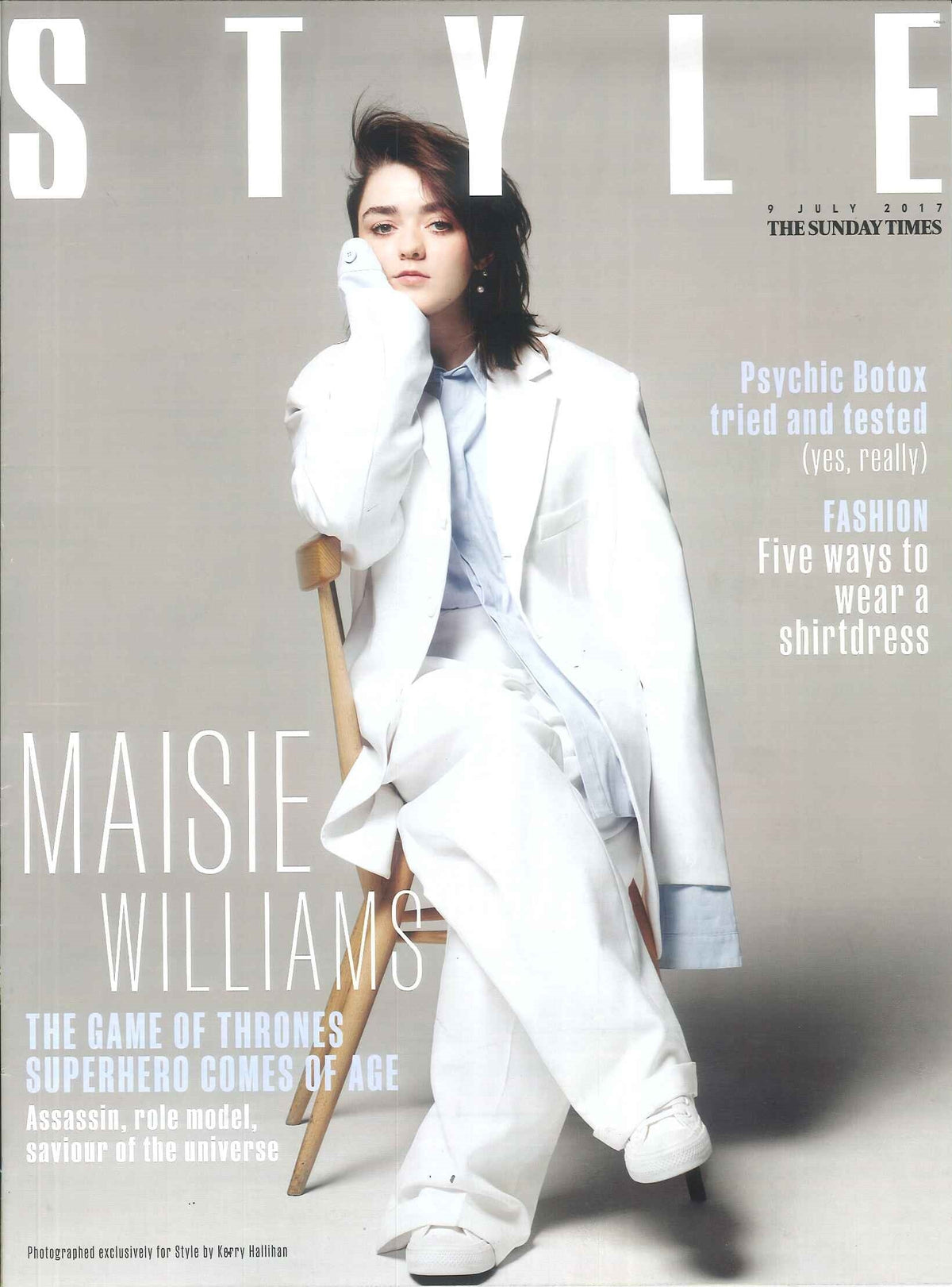 Young woman seated on a wooden chair wearing a loose white blazer, wide-legged trousers, white trainers, and a pale blue shirt