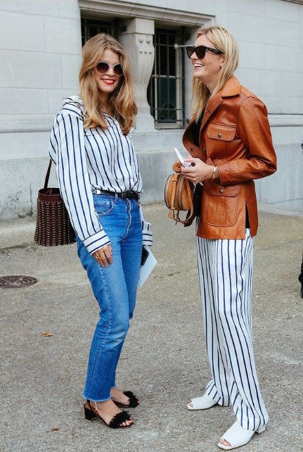 Two women outdoors chatting, one in a white striped blouse and jeans, the other in a brown leather jacket and striped pants