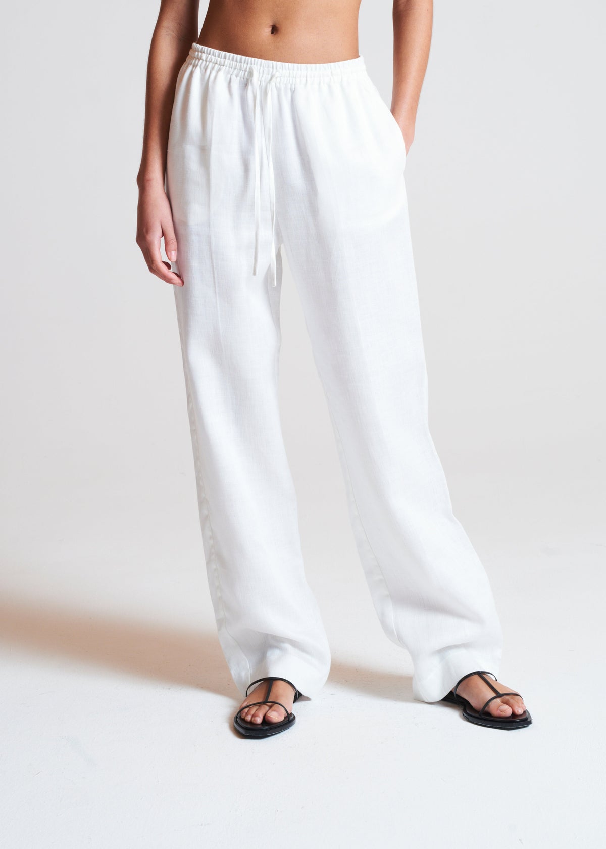 Loose white linen trousers with elastic waistband and drawstring, styled with black thong sandals, shown on model from mid-thigh down