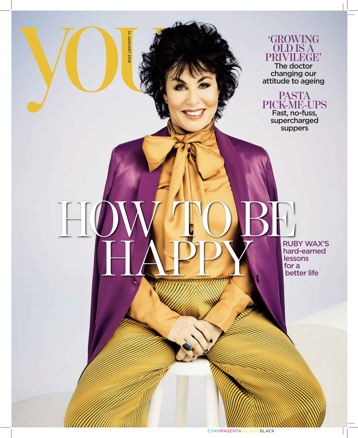 Middle-aged woman seated on white stool wearing mustard blouse with bow, purple blazer, and yellow striped trousers, smiling
