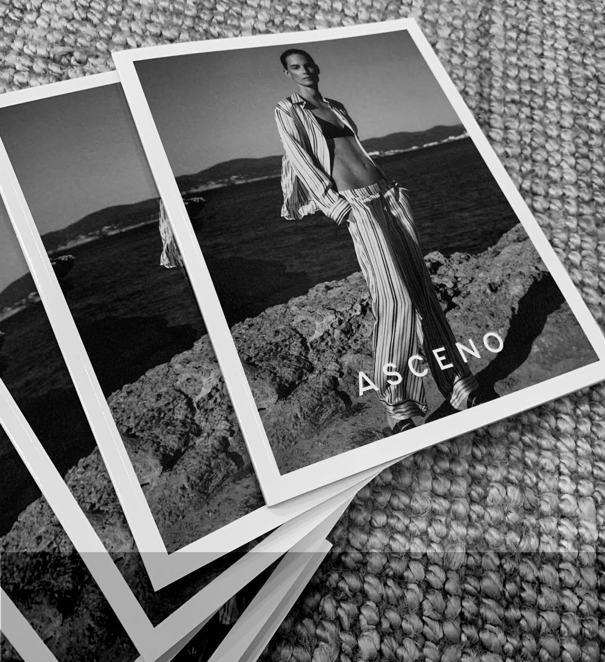 Black and white photo of a model by the sea in striped pyjama pants and an open matching shirt over a dark top, with ASCENO logo