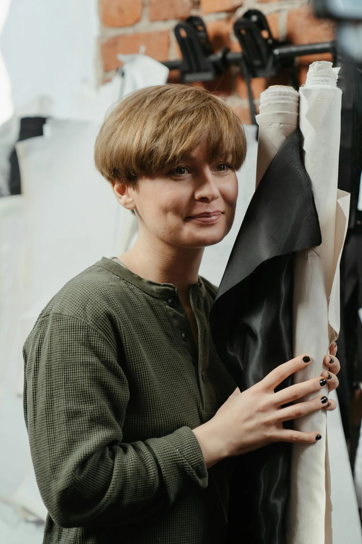 Person in olive green waffle-knit top holding rolls of black, white, and cream fabric in a studio with a red brick wall