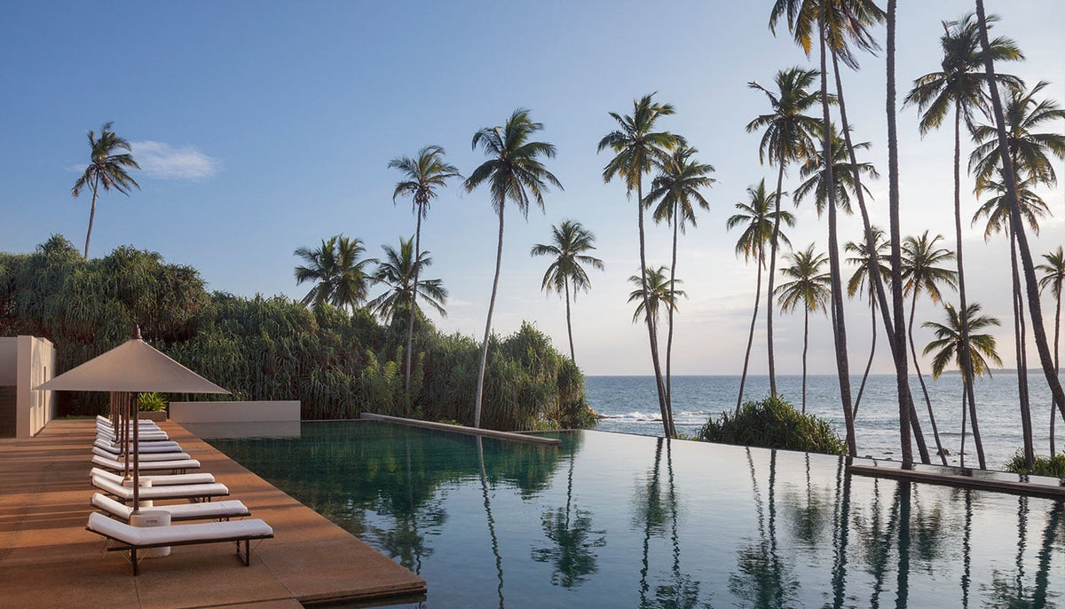 Infinity pool beside white-cushioned loungers and beige umbrellas with tall palm trees and ocean view in natural daylight
