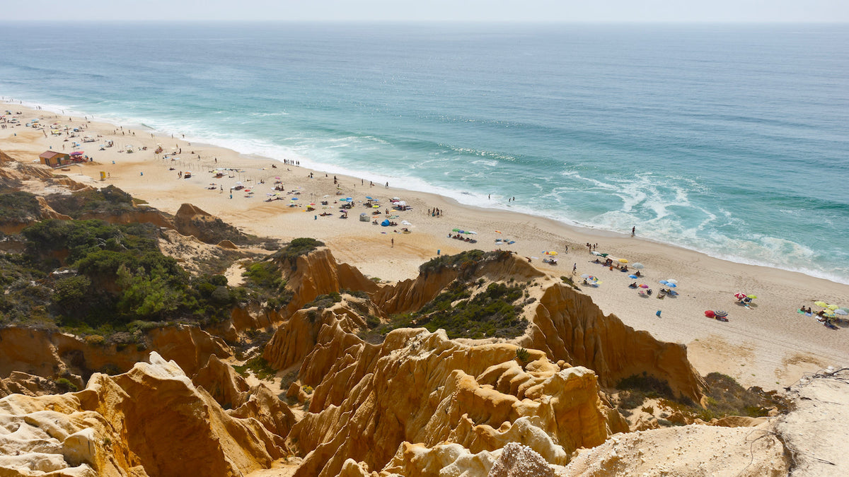 Wide elevated view of a sandy beach with colourful umbrellas below rugged orange cliffs and a calm blue ocean