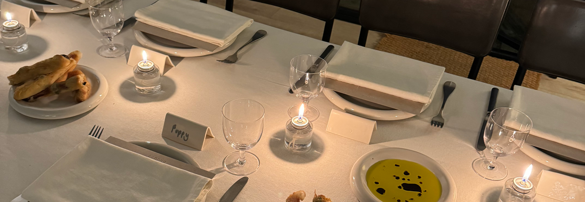 Warmly lit dining table set with white linen, glassware, folded napkins, candles, breadsticks, and olive oil dip