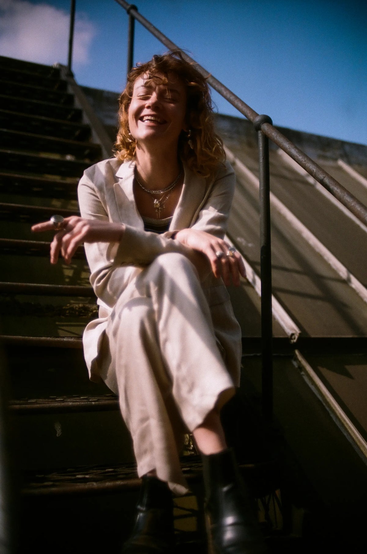 Relaxed woman on outdoor metal stairs wearing a light beige suit, striped top, and black ankle boots smiling with eyes closed