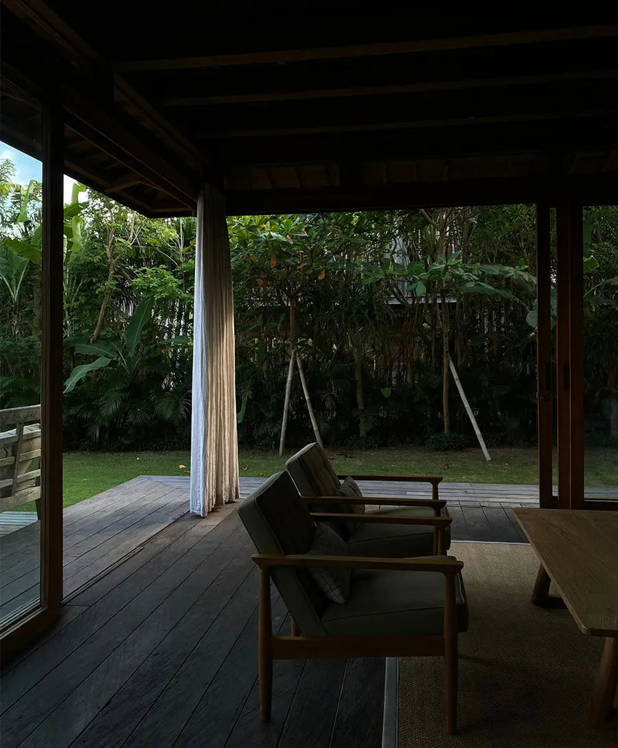Two wooden framed cushioned chairs with pillows face a wooden coffee table on a rug in a dim indoor space opening to a green garden