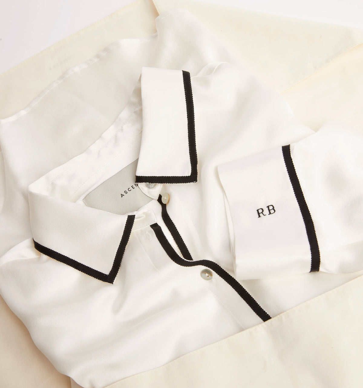 White silk pajama top with black trim on collar, cuffs, and button placket, embroidered with initials 'RB' on cuff