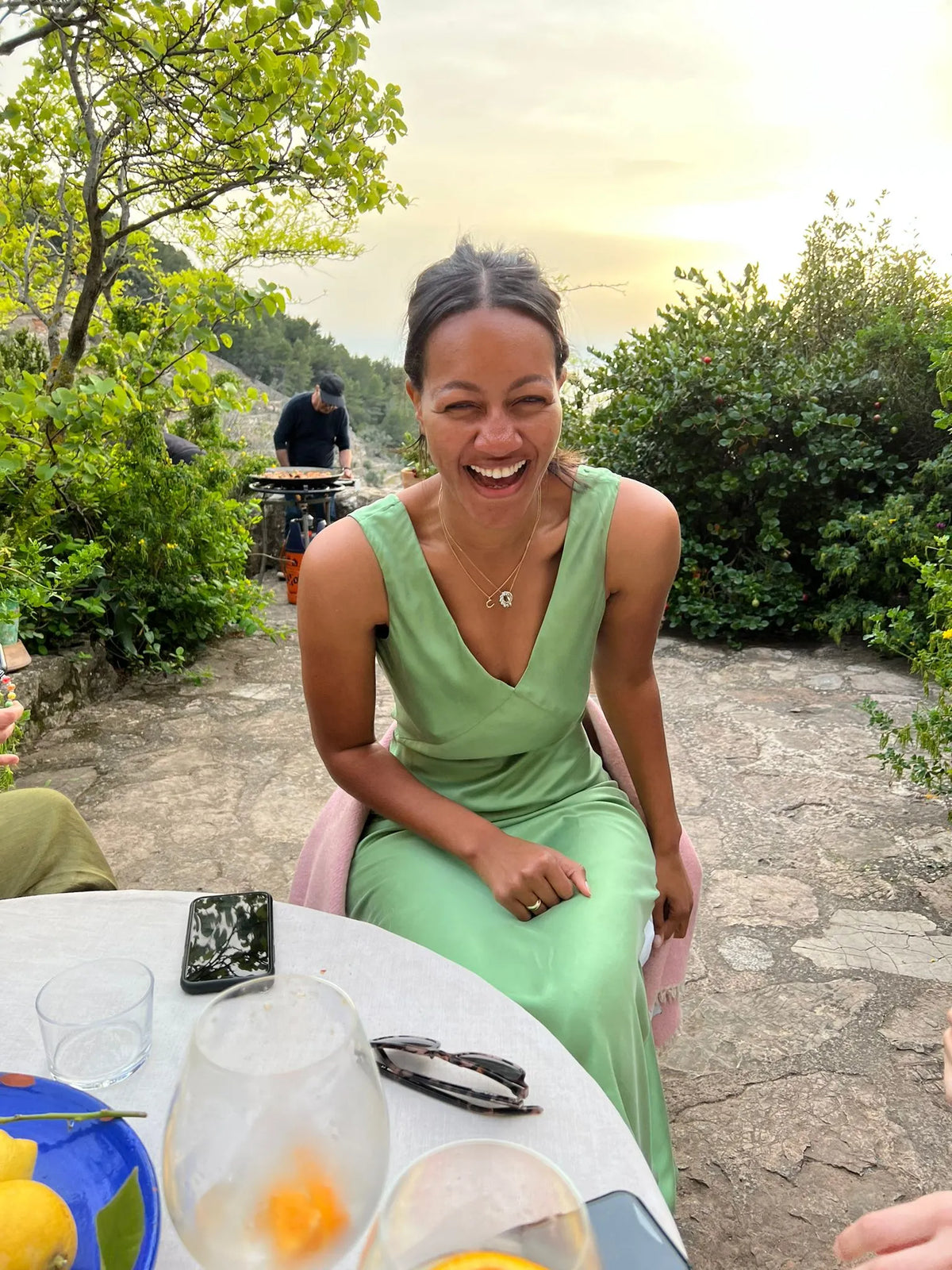 Woman laughing in a sleeveless light green dress seated outdoors on a stone patio with greenery and a barbecue grill behind her