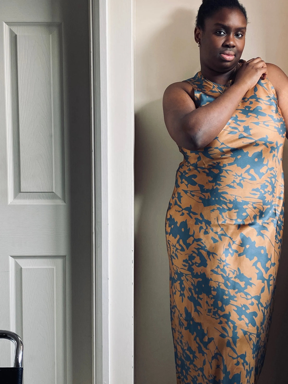 Sleeveless fitted dress with blue botanical print on tan fabric, shown on model standing indoors by a white door