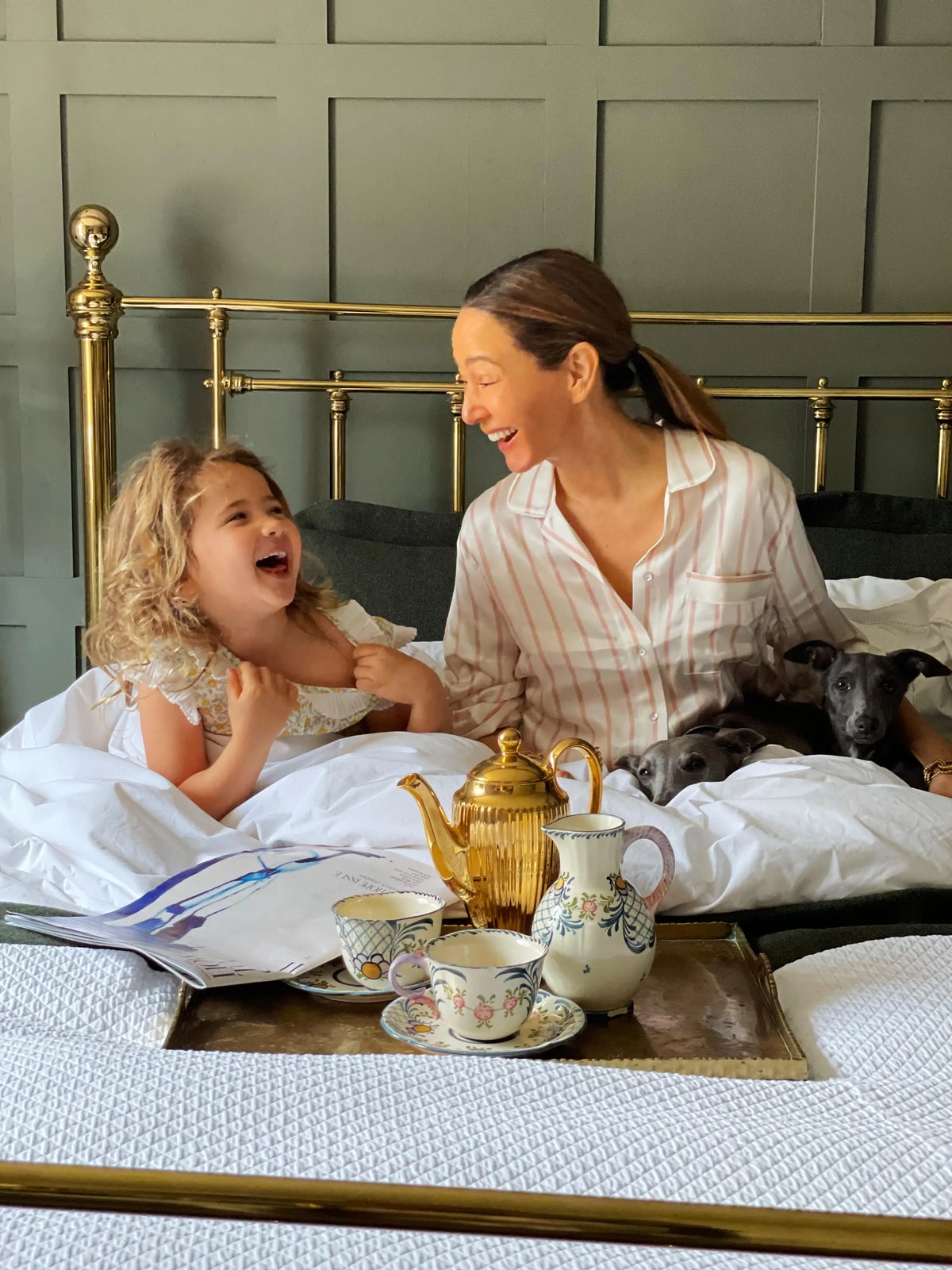 Woman in pale striped Asceno silk pyjamas and child sharing a joyful moment on a brass bed with white linens