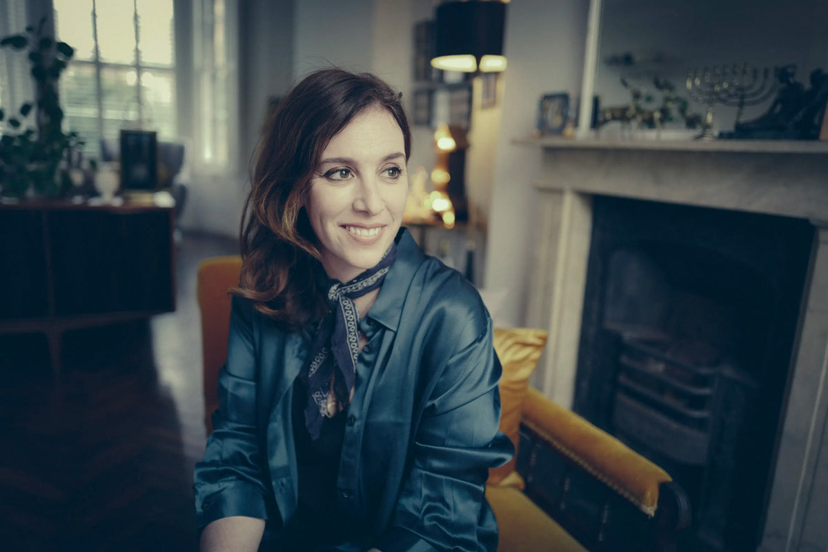 Woman seated on mustard-yellow armchair wearing a dark teal satin blouse and patterned scarf in a softly lit room