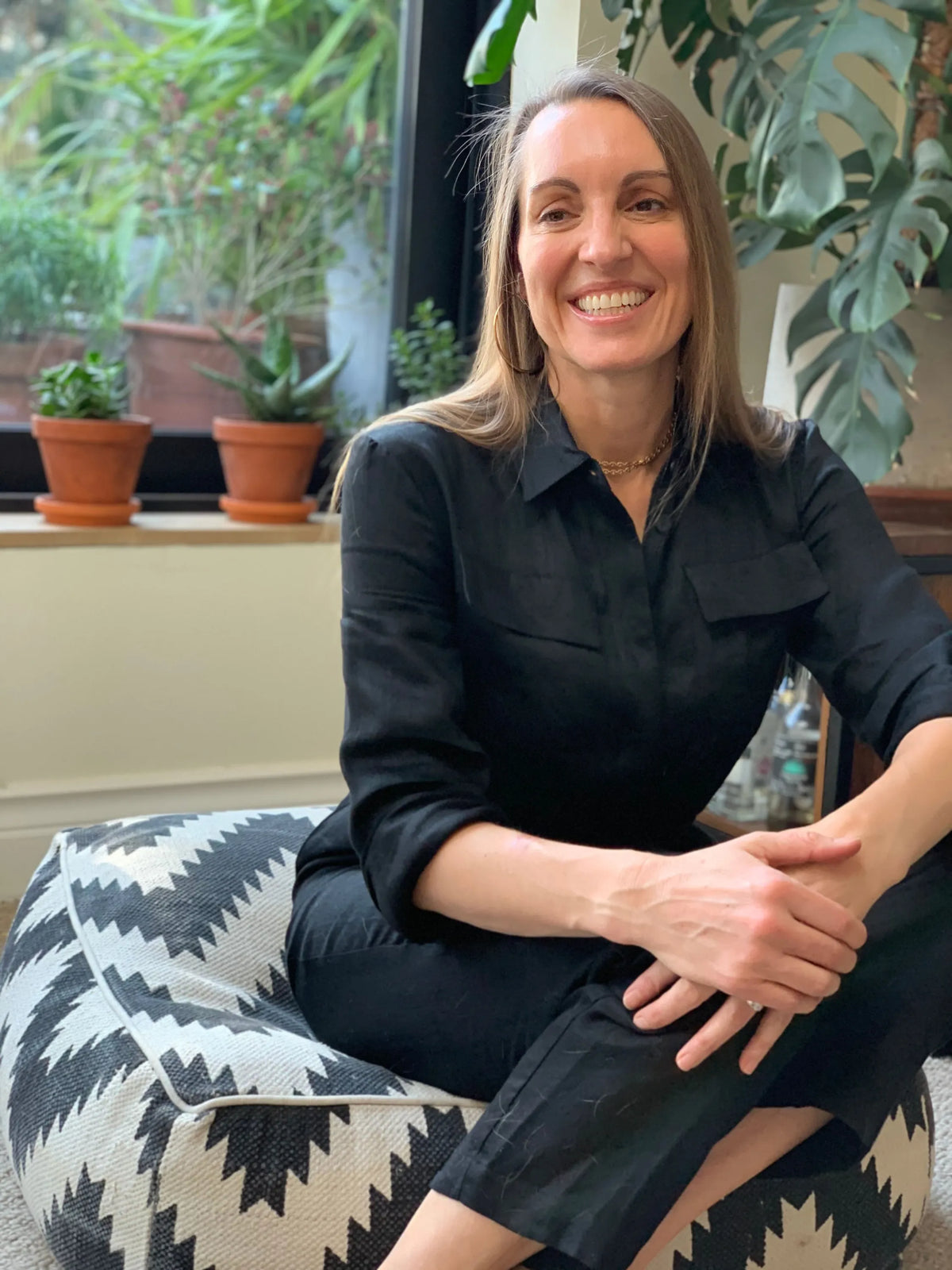 Woman seated on monochrome zigzag cushion wearing relaxed black shirt and trousers, smiling softly by a sunlit window with potted plants