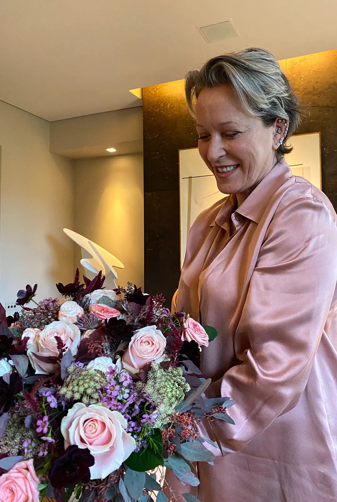 Middle-aged woman in a light pink satin shirt arranging a large bouquet of pink roses and mixed flowers indoors