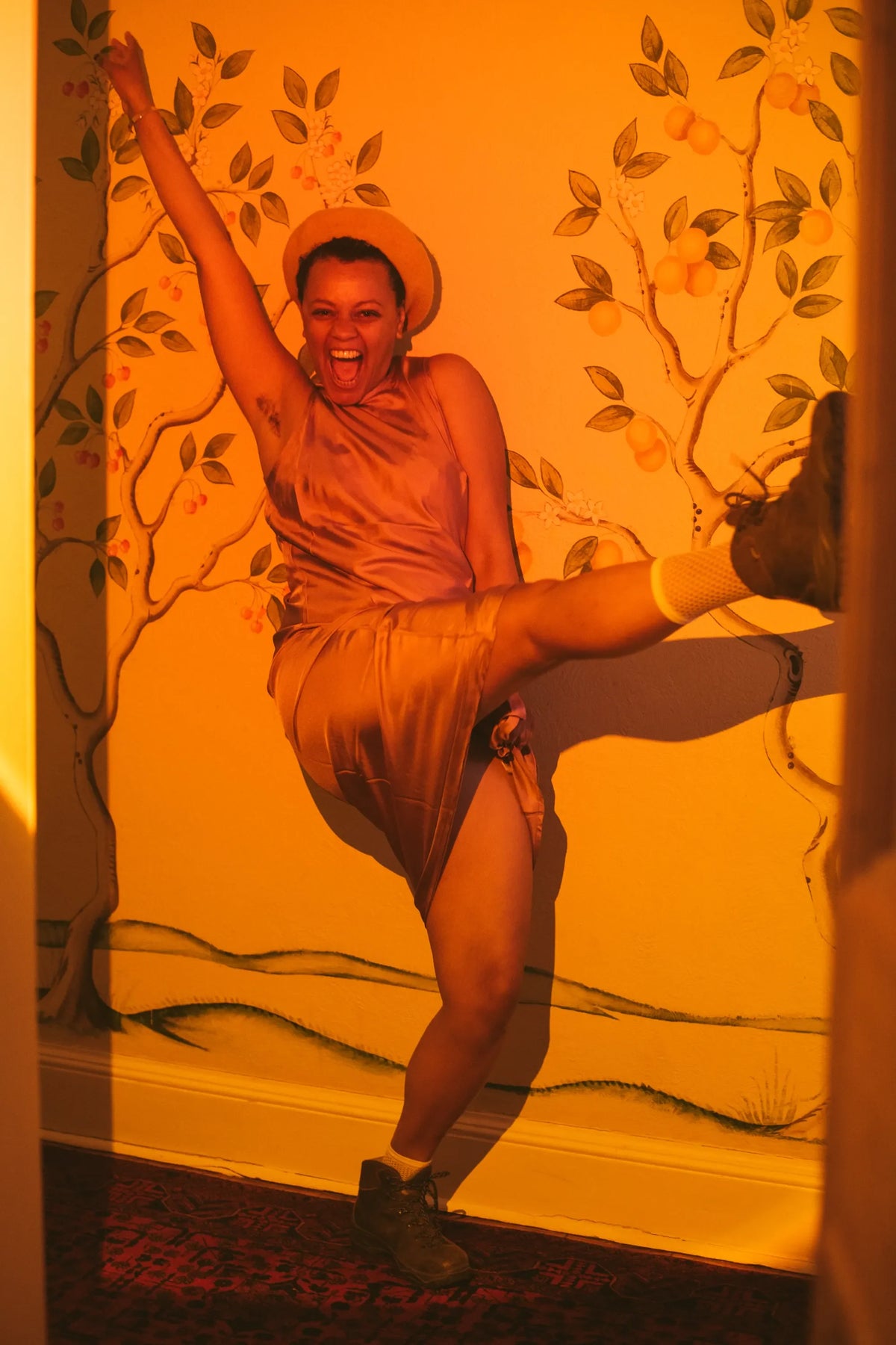 Woman in a pink satin sleeveless dress and beige hat kicking leg high indoors with warm orange light and tree mural background