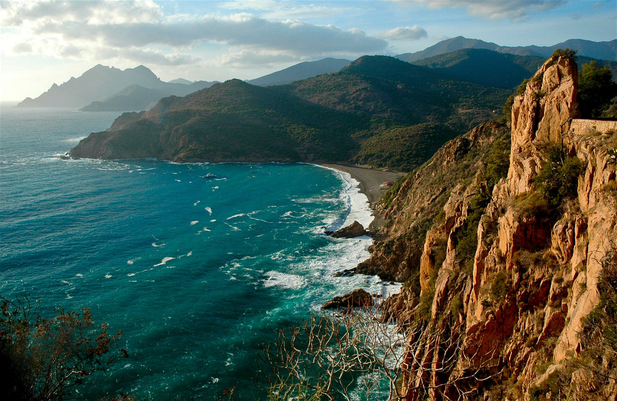 Turquoise sea waves meet a curved beach bordered by reddish cliffs and green shrub-covered hills under a partly cloudy sky
