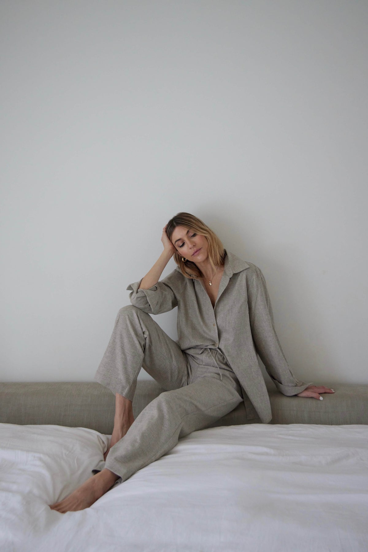 Woman seated on bed edge wearing a relaxed light grey linen shirt and matching trousers in soft natural light