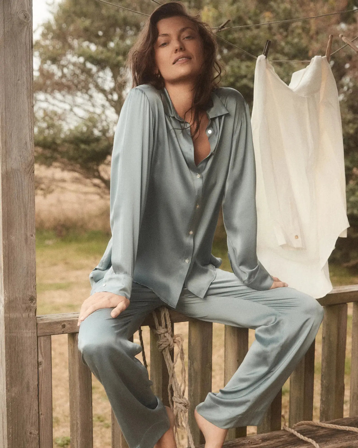 Young woman in relaxed pose wearing light blue-gray satin pyjama set seated barefoot on wooden porch railing outdoors
