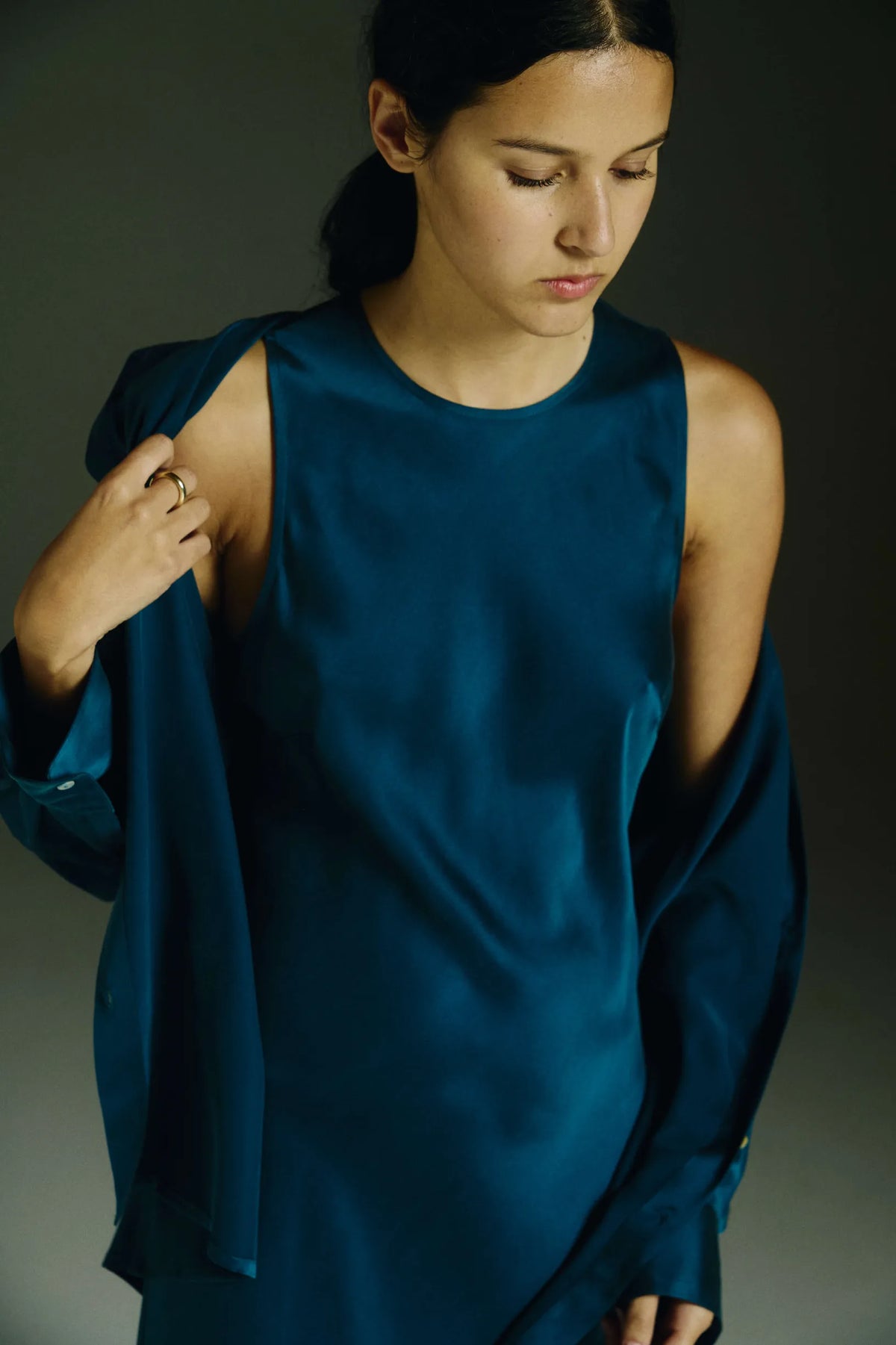 Woman adjusting a dark teal satin sleeveless dress with a matching long-sleeve shirt against a dark background