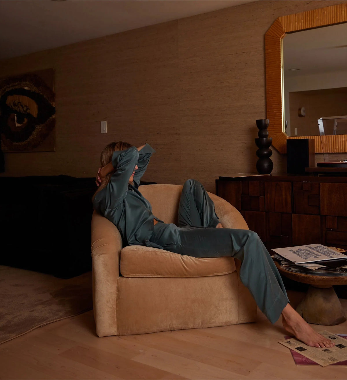 Relaxed woman reclining sideways in dark teal silk pyjamas on a beige armchair in a warmly lit living room