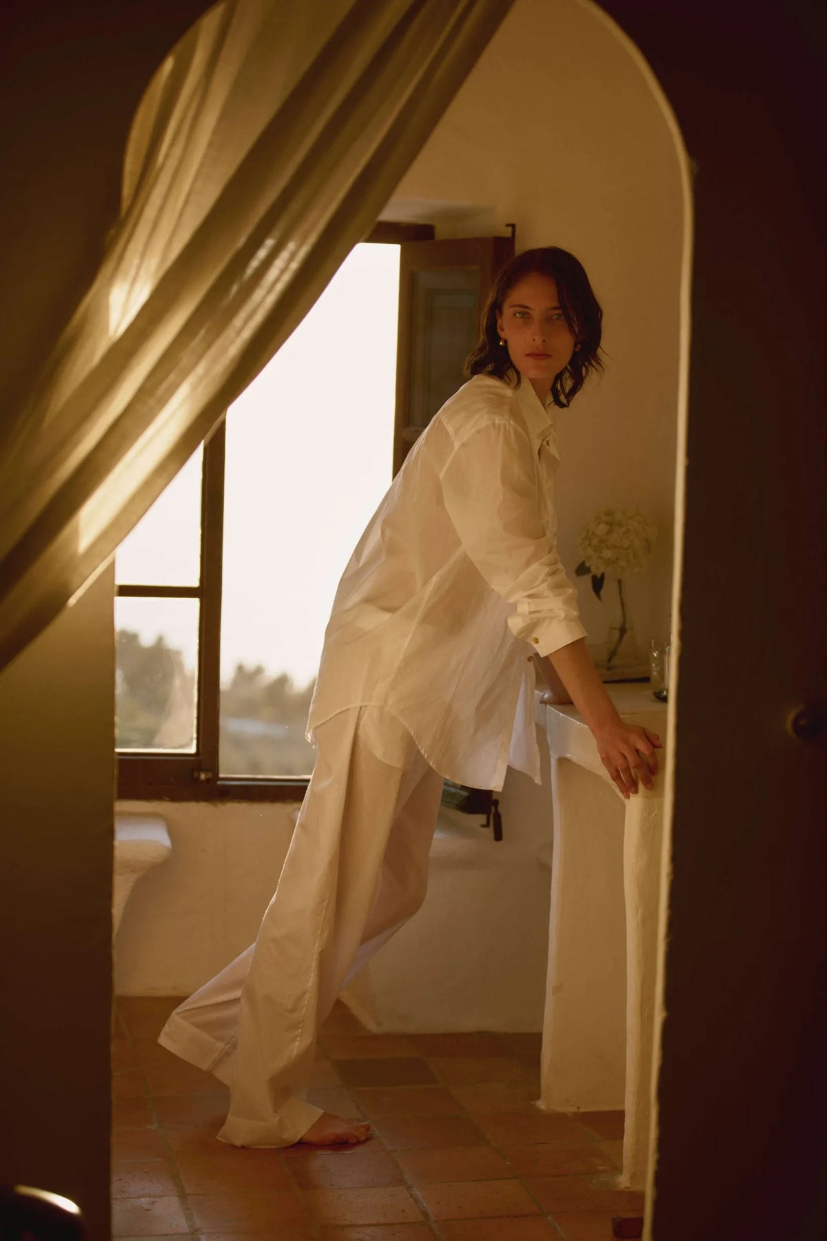 Woman in loose, all-white linen shirt and wide-legged trousers leaning on a white sink in a warmly lit room with terracotta floor tiles