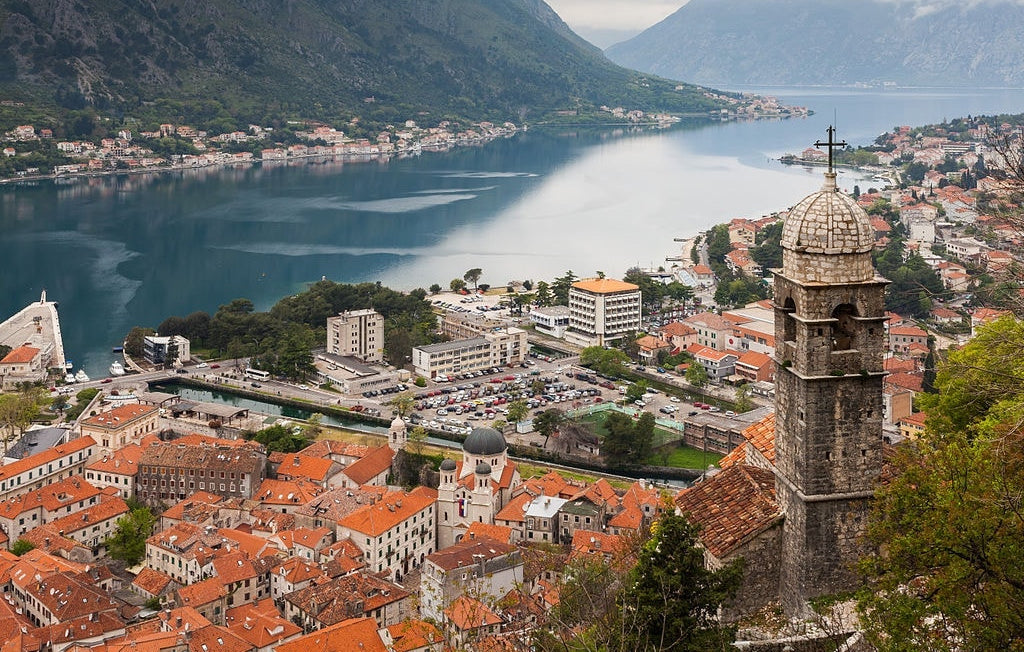 Panoramic view of a coastal town with orange-tiled roofs, a stone bell tower in foreground, calm water and mountains behind