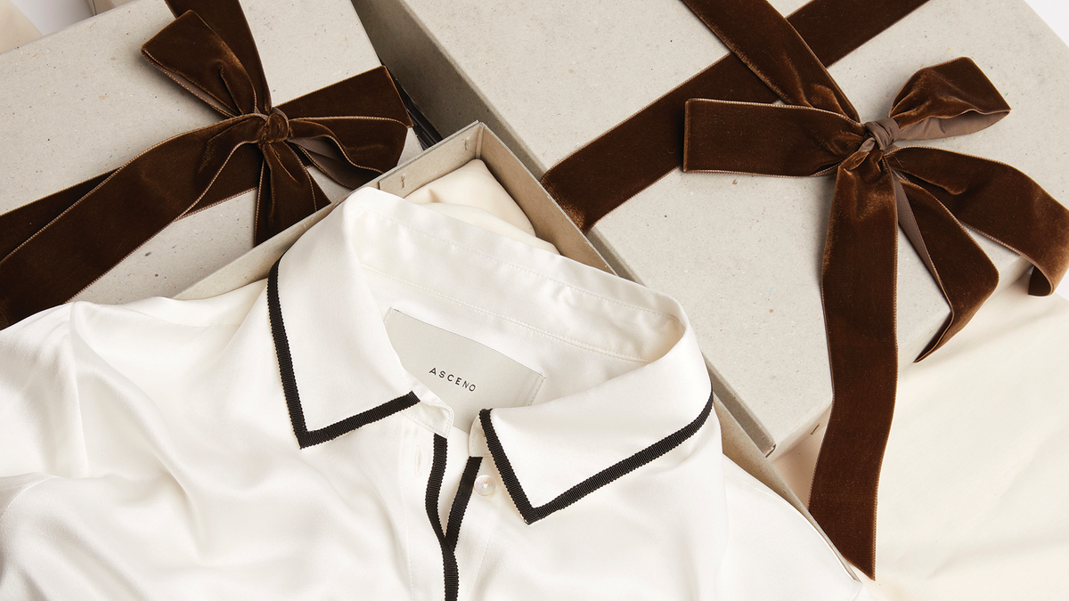 White silk blouse with black edging on collar and button seam, inside open beige gift box next to closed box with brown velvet bow