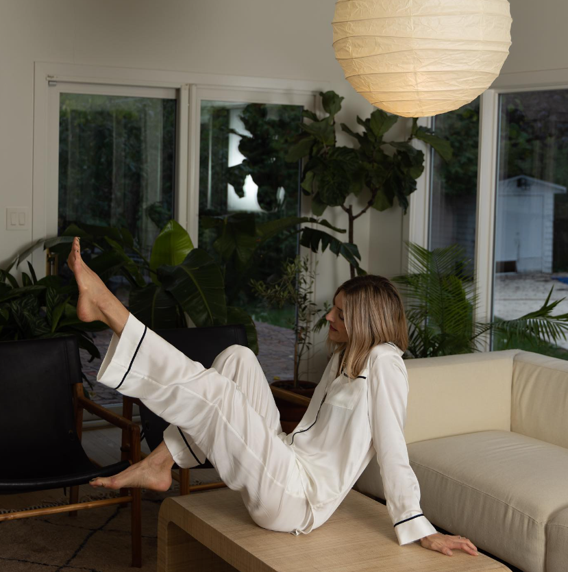 Woman seated on light wood table in white silk pyjamas with black trim, in a modern living room with plants