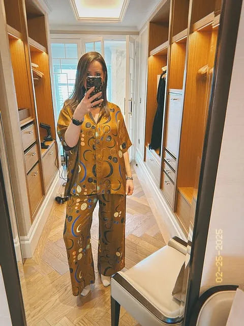 Woman wearing brown silk pyjamas with blue and white abstract circular patterns, taking mirror selfie in wooden walk-in closet