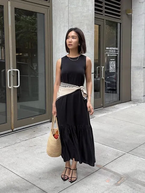 Elegant woman in a sleeveless black maxi dress with a light scarf tied at the waist, carrying a woven straw tote on an urban sidewalk