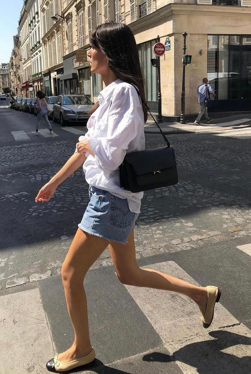 Woman walking on cobblestone street in white blouse, denim shorts, beige flats with black toes, and carrying black handbag