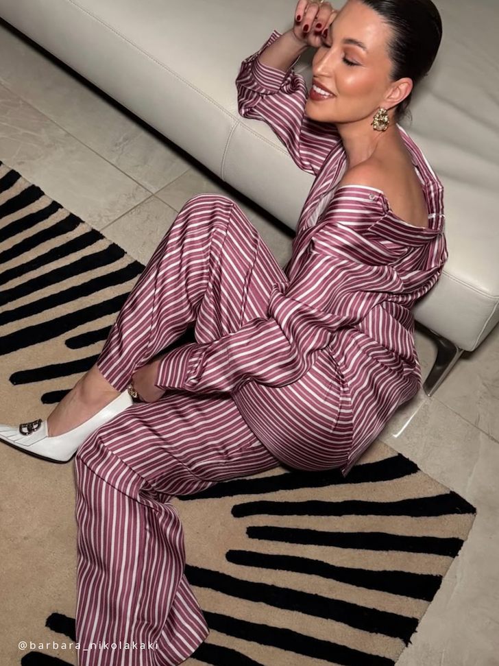 Woman seated on floor in maroon and white striped silk pyjamas with off-shoulder top and white heels, beside cream leather sofa