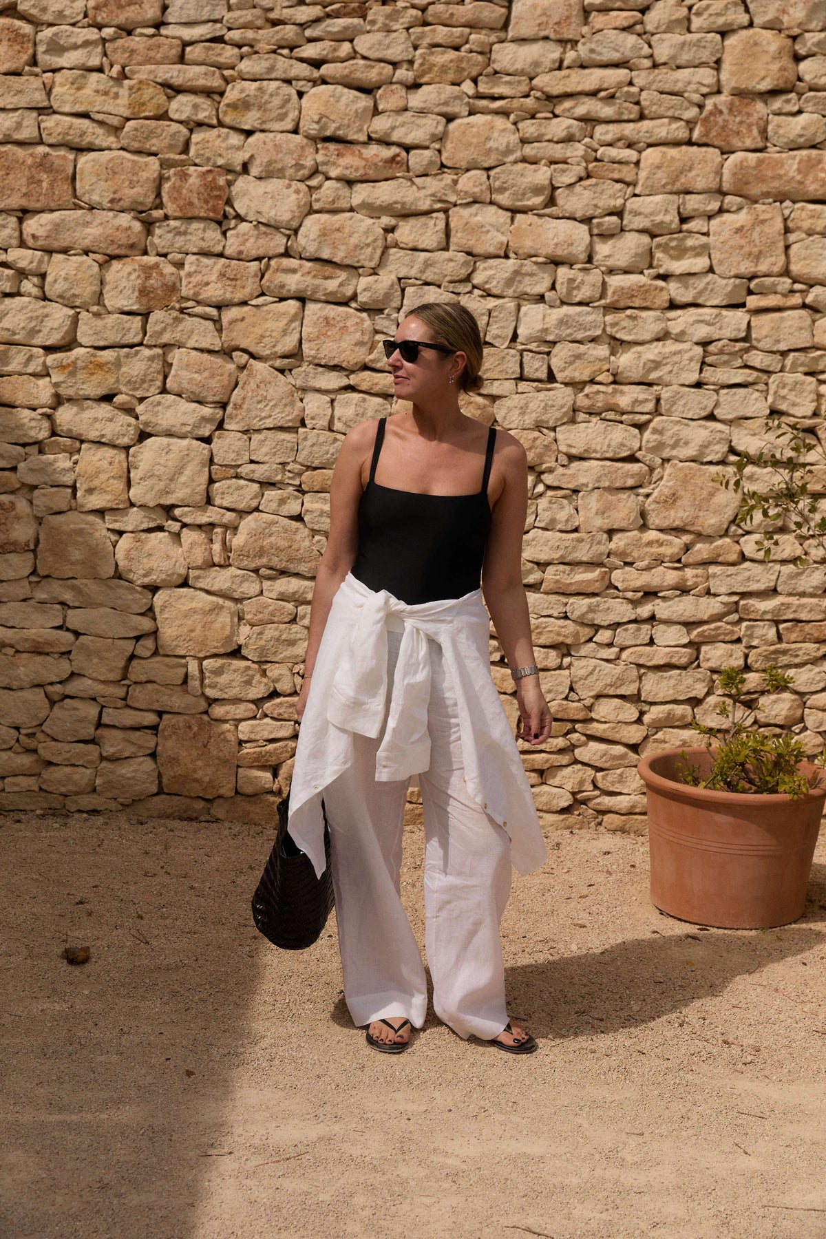 Woman in a black sleeveless top, white linen pants, and a white shirt tied at the waist holding a black woven tote by a stone wall