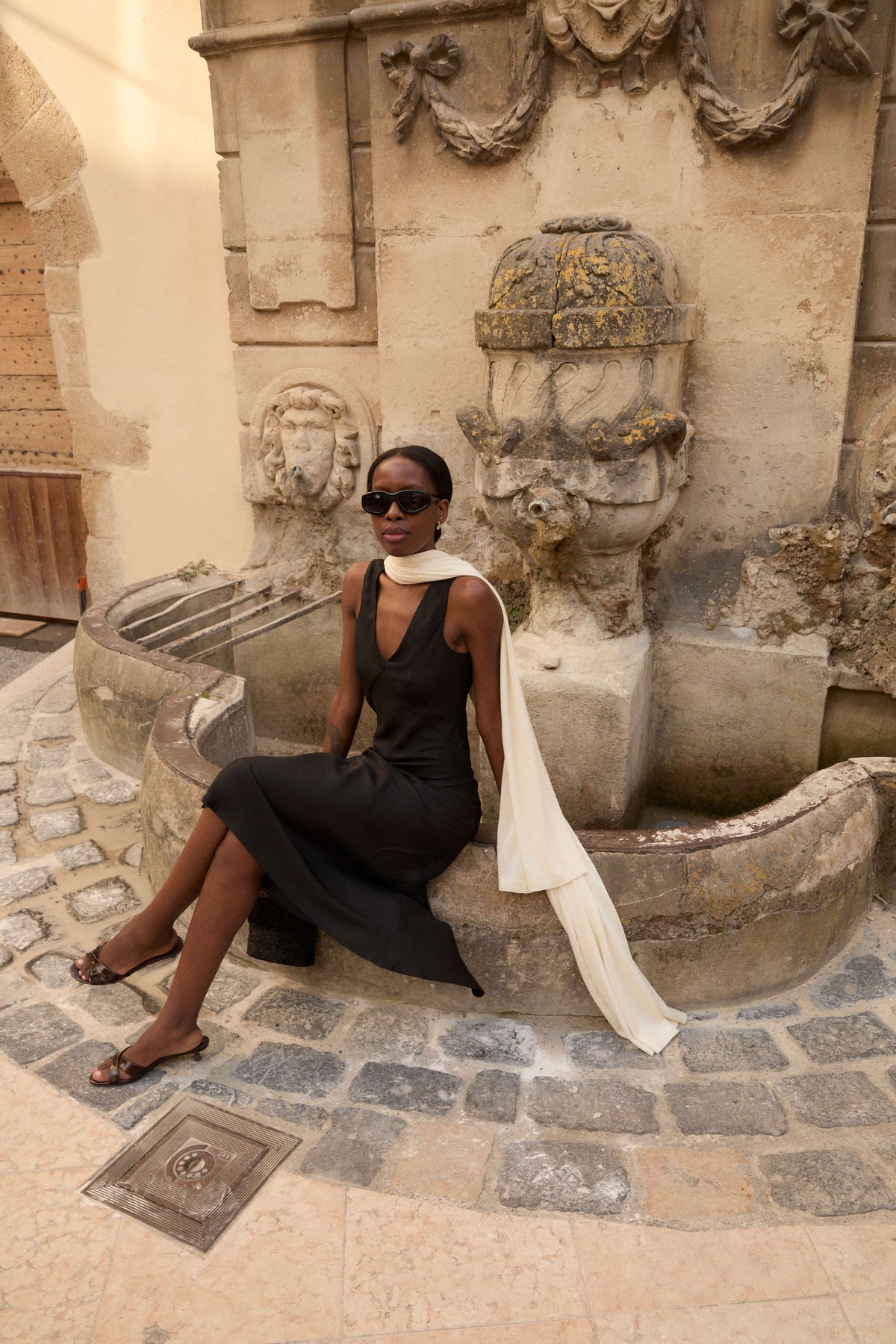 Elegant woman seated on stone fountain edge wearing a black sleeveless dress, white scarf, brown sandals, and dark sunglasses