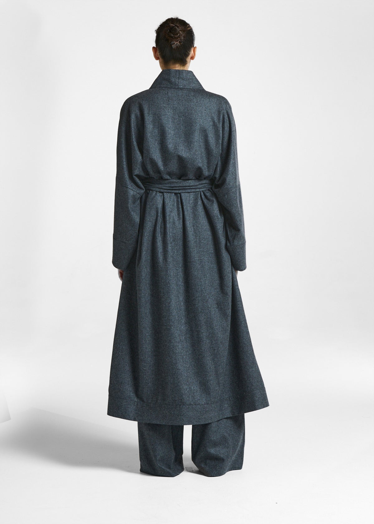 Back view of a woman wearing a belted dark grey long coat with wide-legged matching trousers against a white background