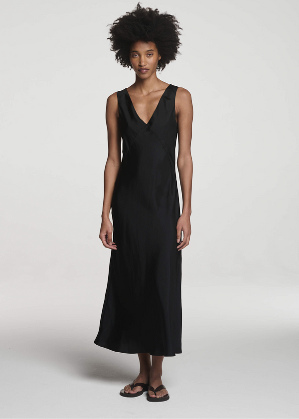 Woman wearing a sleeveless black silk maxi dress with a V-neckline and black sandals, standing against a light grey background