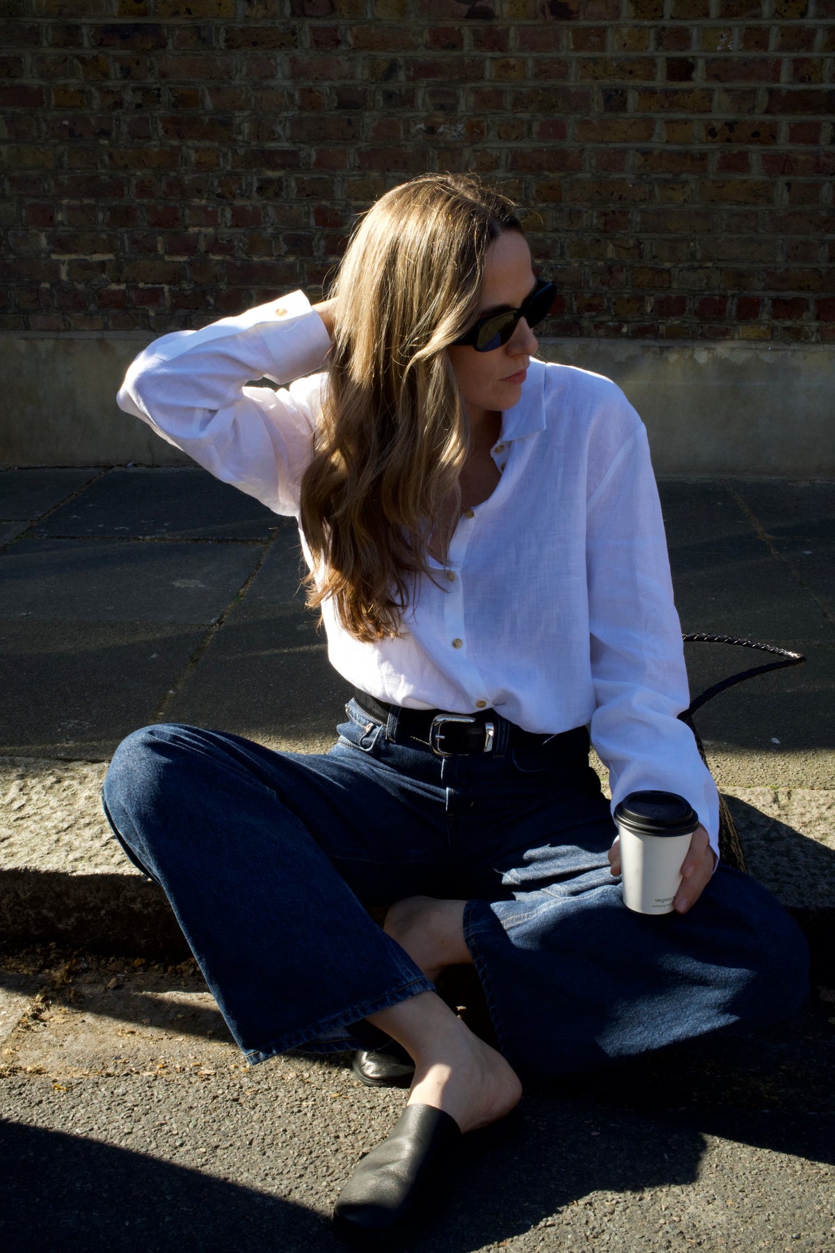 Woman in white linen shirt and dark wide-leg jeans sitting outdoors on a stone curb holding a coffee cup