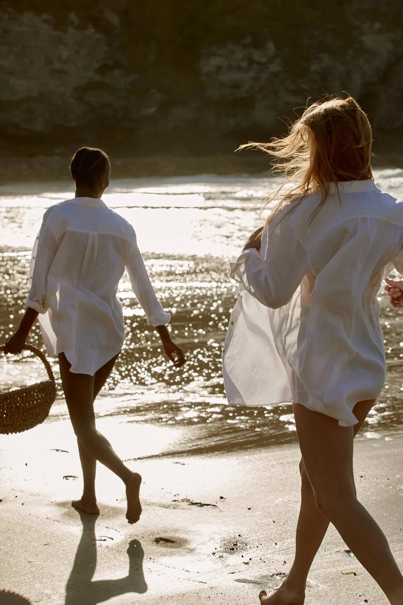 {"image_loaded": true, "load_issue": null, "description": "The image shows two women walking barefoot along a sandy beach with gentle waves reflecting sunlight in the background. Both are wearing loose, long white shirts with the sleeves rolled up; one woman has light hair that is blowing in the wind, and the other has darker skin with braided hair and carries a woven basket. The scene is backlit, creating silhouettes and shimmering reflections on the wet sand.", "people": {"count": 2, "roles": ["beachgoers